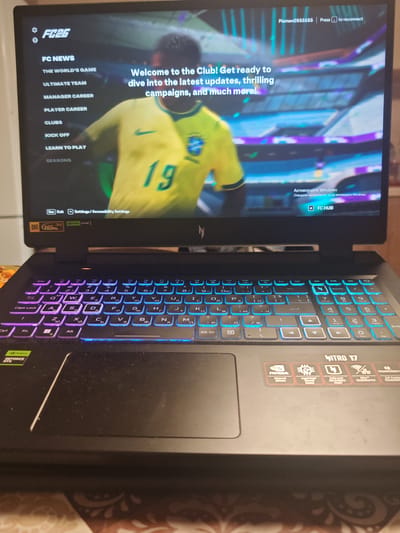 The image shows a laptop screen displaying the FC 24 video game menu. The menu options include "FC NEWS", "THE WORLD'S GAME", "ULTIMATE TEAM", "MANAGER CAREER", "PLAYER CAREER", "CLUBS", "KICK OFF", "LEARN TO PLAY", and "SEASONS". The game features a player in a yellow and green football kit with the number 19 on the back. The laptop itself has a backlit keyboard with various keys visible, including "Esc", "Fn", "Ctrl", "Alt", and function keys. There are also stickers on the laptop, including "NVIDIA GEFORCE RTX" and "NINTRO 17". The background of the laptop screen shows a stadium with bright lights and purple and blue accents. The overall setting appears to be indoors, likely a room within a home or office. There is no visible indication of the city of Podvis, Bulgaria, in the image itself.
