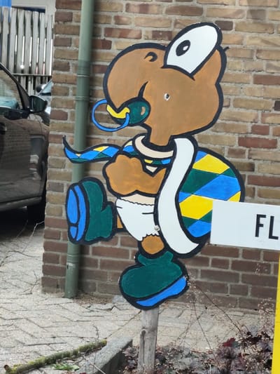 A cartoon turtle figure painted on a wooden cutout stands outdoors, leaning against a brick wall. The turtle is depicted with brown skin, large eyes, a blue pacifier in its mouth, and a green, yellow, and blue patterned shell. It is wearing a white diaper and green shoes with blue accents. A white sign with the letters "FL" is partially visible in front of the turtle. The turtle figure is mounted on a wooden post. The environment includes a brick wall, paving stones, a drainpipe, a parked car on the left, and some vegetation. The overall scene suggests an outdoor decorative element, possibly in a garden or public space in the city of Schaijk, Netherlands.