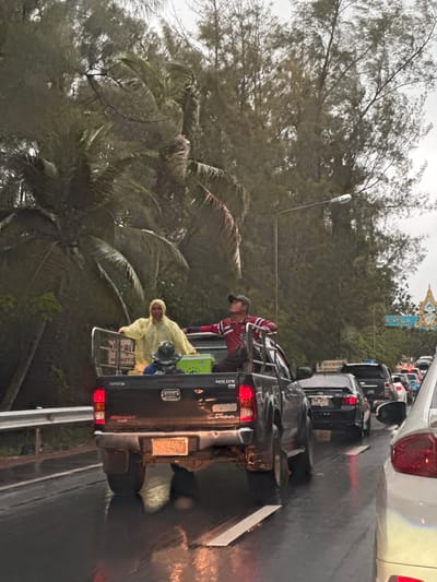 This image depicts a traffic scene on a wet road, likely in Baan That Chatchai, Thailand, due to the presence of tropical vegetation and a roadside shrine with a portrait of a royal figure. A black Toyota Hilux pickup truck is the central focus. Two men are visible in the truck bed. One man, wearing a bright yellow raincoat, is leaning on the truck's side rail, facing left. The other man, wearing a red and white striped shirt and a dark baseball cap, is standing and looking upwards, also in the truck bed. A green cooler is also present in the truck bed. Several other vehicles are on the road, indicating a traffic jam, and the road surface is reflective due to rain. Lush green trees and palm fronds line the sides of the road. A street lamp is visible overhead. The sky is overcast. The license plate on the pickup truck is partially obscured but appears to read "UB 4057".