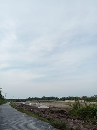 The image depicts an overcast sky above a rural scene in Teluk Kenidai, Indonesia. In the foreground, a paved road with distinct horizontal ribbing runs diagonally from the lower left to the mid-left of the frame. To the right of the road is an area of disturbed earth, with mounds of sand and piles of dark soil. A small yellow excavator is visible in the mid-ground on the construction site. A line of palm trees forms a distant treeline, separating the construction area from the sky. A power pole with wires is situated on the left side of the road, and two motorcycles are seen on the road in the distance. The overall environment appears to be a developing area, possibly undergoing construction or land preparation.