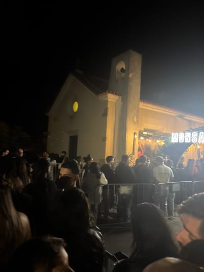 The image shows a nighttime scene outside a building, likely a restaurant or bar based on the illuminated signage that reads "MONSA." A crowd of people is gathered, some of whom are behind metal barriers, suggesting a queue or waiting area. The building itself appears to be an older structure with architectural details like a bell tower or small arched window. The lighting is a mix of artificial light from the signage and building, contrasting against the darker night sky. The location is indicated as Amadora, Portugal, based on the establishment's location.