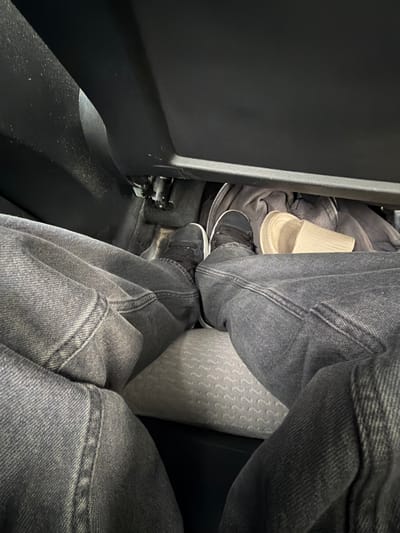 This image provides a low-angle, close-up view of a person's legs and feet inside a vehicle, likely a car, as indicated by the presence of a seat and car interior elements. The person is wearing dark denim jeans and dark sneakers. A pair of light-colored, slide-style slippers are visible tucked away in the footwell alongside a dark grey hooded sweatshirt. The lighting suggests the interior of the vehicle is dimly lit, possibly at night or in a shaded area. The scene appears to be domestic or personal, with no indication of a public or commercial setting within Vallensbæk Landsby, Denmark.