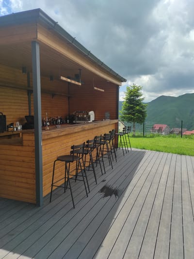 An outdoor wooden bar area is captured in Gudauri, Georgia, under a cloudy sky. The bar itself is constructed from light-colored wood, with a smooth countertop displaying various bottles of liquor, a coffee machine, and other bar essentials. In front of the bar, five black metal stools with circular seats are arranged neatly on a grey wooden deck. A portion of the deck is wet, possibly from recent rain. To the left of the bar, a speaker and a small black laptop are visible.
Behind the bar, the wooden structure continues, with a slanted roof supported by a grey pillar. The background reveals a lush green mountainous landscape, characteristic of Gudauri. A tall, mature evergreen tree stands to the right of the bar, and further in the distance, several houses with red roofs are nestled among the hills. A metal fence separates the grassy area from the more distant terrain. The overall scene suggests a serene and picturesque setting for an outdoor establishment.