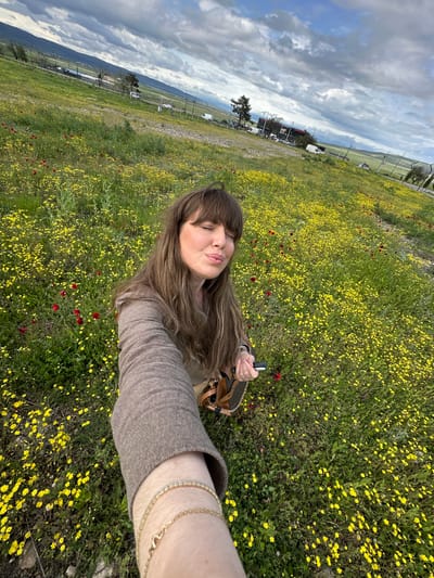 A selfie depicts a woman with long, brown hair, and bangs, pouting her lips. She is positioned in a field filled with yellow wildflowers and sporadic red poppies. The photo is taken from a low angle, with the camera held at arm's length, capturing her face and arm in the foreground. She is wearing a brown jacket and at least one gold bracelet on her wrist, and holding a small object. The background includes a cloudy sky, distant mountains, and what appears to be a roadside area with utility poles, buildings, and parked vehicles. This scene could potentially be located near Marneuli, Georgia, given the landscape and vegetation.