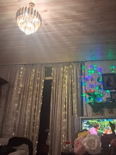 An interior scene in Lanchkhuti, Georgia, depicts a decorated residential room, likely a living area, illuminated by artificial light. A large, multi-tiered chandelier with wooden and crystalline elements hangs from a light-colored, horizontal wooden plank ceiling in the upper center. The left and central portions of the image are dominated by floor-length, light-beige or gold-patterned curtains, strung with numerous small, warm white fairy lights. A dark window is partially visible between the curtains. To the right, a wall is adorned with multicolored (blue, green, red, purple) string lights, multiple framed religious icons and photographs, and what appear to be calendars or posters below a small shelf. The shelf holds a vase containing red and orange artificial flowers. In the lower right, an older model cathode-ray tube television displays a vibrant outdoor scene featuring two individuals, one in a pink shirt and another with red hair wearing what appears to be a virtual reality headset. In the immediate foreground, on a surface in front of the television, a collection of artificial roses in shades of pink, white, yellow, and red is arranged next to a plastic bottle of a dark carbonated beverage with a red cap. A dark-colored upholstered furniture piece with a white covering is partially visible in the lower left. No individuals are present within the depicted room itself.
