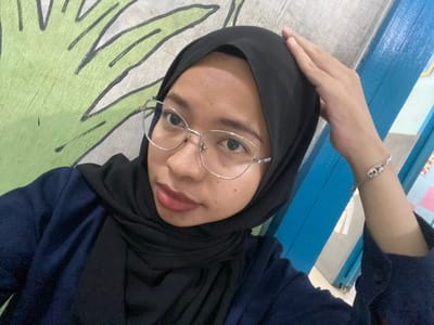 In Tanjung Gading, Indonesia, a young woman wearing a black hijab and glasses is taking a selfie. She is wearing a navy blue jacket over a black top. Her right hand is touching her head. A portion of a wall mural with green leaf designs is visible in the background, as well as a blue wooden structure. A silver bracelet is on her left wrist.
