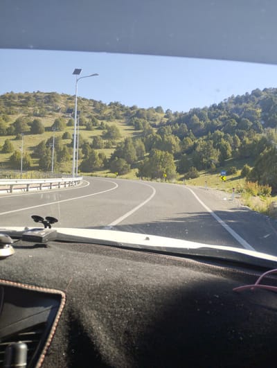 The image shows a view from inside a vehicle looking out at a winding road in a hilly, sparsely vegetated area under a clear blue sky. The road has white lane markings and a guardrail on the left side. Several streetlights with solar panels are visible along the road, as well as yellow directional signs. The foreground of the image shows the interior of the vehicle, including the dashboard and windshield, with a device mounted on the dashboard. The vegetation consists of scattered trees and shrubs on the hillsides. The scene appears to be a rural or semi-rural setting. It is not possible to determine the exact location within Myk, Uzbekistan from the image.
