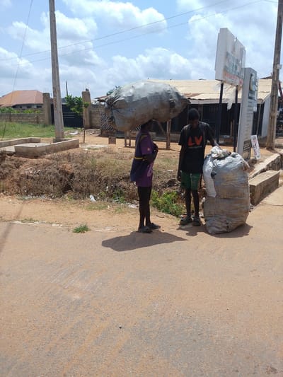 In Iseyin, Nigeria, two young men are pictured standing on a dirt road next to large, wrapped bundles. One man, wearing a purple shirt and dark pants, carries a large, grey, sack-like bundle on his head. The other man, in a black t-shirt with an orange "LV LOUIS VUITTON" logo and green shorts, stands beside a tall, cylindrical bundle wrapped in a white and grey material. They are positioned near a street with a concrete structure and a sparse, overgrown embankment filled with debris. In the background, corrugated metal roofs of buildings and a utility pole are visible under a cloudy sky.