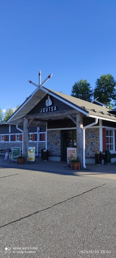 The image shows the exterior of a building with the word "JOUTSA" prominently displayed above the entrance. The building has a rustic facade with stone and dark wood siding, and red-shuttered windows. Three white and red decorative elements are mounted on the roof peak. Several signs are placed outside the building, including one that appears to be an advertisement. Two dark trash receptacles are located near the entrance. The building is situated on an asphalted area, possibly a parking lot, with a clear blue sky overhead. The overall setting suggests a commercial establishment or public facility in Joutsa, Finland.