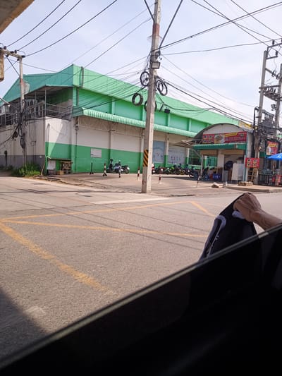 The image depicts a street scene in Hua Hin City Municipality, Thailand. The camera appears to be inside a vehicle, focusing on the intersection. The road features yellow crosswalk markings. On the opposite side of the road stands a large building with a green roof and green and white striped walls. Several motorcycles are parked in front of the building. Power lines are visible overhead. A hand holding a black and white garment is visible within the vehicle. A food stall is situated to the right of the building.