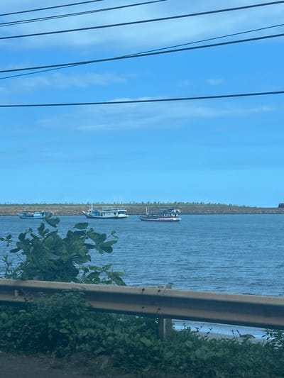 In Borongloe, Indonesia, a body of water, likely a strait or bay, is visible under a blue sky with scattered clouds. Three boats are sailing on the water. The boat on the far left is blue and white, the middle boat is predominantly white with blue accents, and the boat on the right is red and white. A breakwater or seawall made of rocks lines the far shore, with sparse vegetation growing on it. In the foreground, a rusty metal railing runs horizontally, partially obscured by lush green foliage.