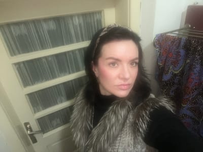 A woman with dark hair and a patterned headband is pictured indoors. She wears a black turtleneck sweater under a gray faux fur vest. A door with multiple glass panes is behind her. To the right of the woman, a garment rack holds a patterned blue and red fabric. The location in Smederevska Palanka Municipality, Serbia is unknown from the visual information provided.