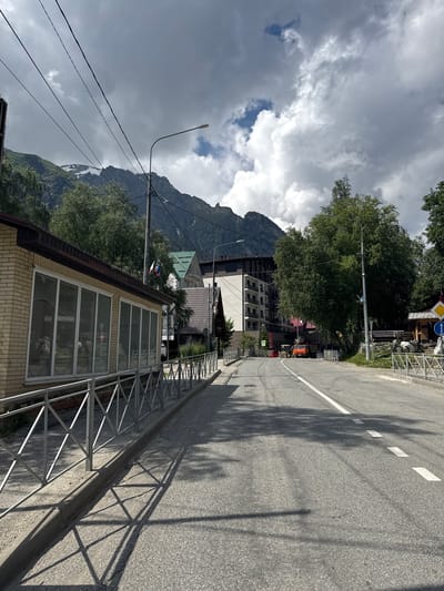 This is a landscape orientation, eye-level, outdoor shot of a road with buildings and a mountain in the background. The road appears to be in a mountainous region, possibly a resort town. The road is paved and has a white dashed line marking the lanes. There is a metal fence along the side of the road, separating it from a building. The building is made of brick and has large windows. In the background, there are several other buildings, trees, and a large mountain. The sky is cloudy, but there are also some patches of blue. The overall impression is one of a scenic, possibly tourist, location.

Specific details include a street light on the left side of the road, and a yellow and blue road sign on the right side of the road. There are also some power lines visible. The time of day appears to be daytime, given the shadows cast by the trees and fence. The weather appears to be partly cloudy.

The location context suggests Dombay, Russia, which is known for its mountains and ski resorts. This image captures the essence of such a location, with its combination of natural beauty and man-made infrastructure. The presence of buildings and a road suggests that this is a developed area, likely catering to tourists. The overall mood of the image is calm and serene, with a sense of natural grandeur.