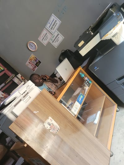 The image shows a cluttered interior scene, likely a small business or office space in Bokkos, Nigeria. A young Black man, wearing a plaid shirt over a light blue t-shirt, is the central subject. He is looking towards the camera and holding a mobile phone to his ear, suggesting he is engaged in a phone conversation.
Various objects are visible, including a large office machine, possibly a printer or copier, to the left. A wooden display cabinet with glass doors contains various items, including what appears to be stationery and a bottle of liquid. A wooden desk or countertop occupies the right side of the frame, with some papers and a torn sticker on its surface. Several small printed signs with phone numbers and company names like "MONIEPOINT," "O OPAY," and "URA BANK" are affixed to a grey wall in the background. A small round clock is also mounted on the wall. In the lower part of the image, a stack of magazines and other printed materials are visible.
The lighting suggests it might be daytime, but no specific time can be determined from the clock face. The overall impression is of a busy, functional workspace with items that indicate financial services or a printing business. The man's expression appears engaged and perhaps slightly tired, consistent with someone working in such an environment.