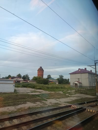 The image, taken from a moving train, shows a landscape with a prominent red brick water tower. The water tower is circular with a conical roof and several arched windows. To the left of the water tower, there are several low-rise buildings, one of which appears to be made of white brick with a grey roof. To the right of the water tower, a two-story building with a red roof and light grey walls is visible. The area surrounding the buildings is overgrown with grass and some trees. In the foreground, train tracks are clearly visible, indicating the perspective of the photographer. The sky is cloudy and a pale blue. The water tower is a distinctive landmark in Miloslavskoye, Russia.