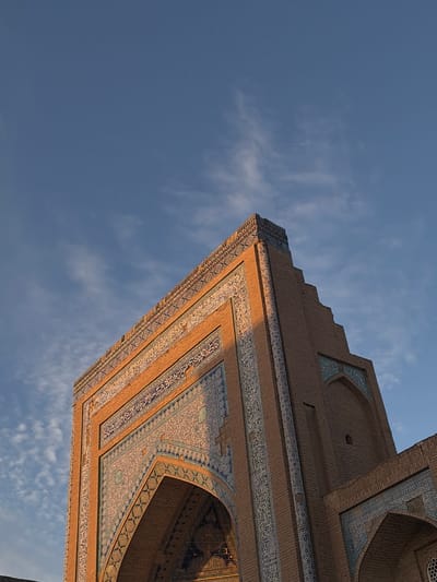 This is a low-angle, full-shot photograph of the western facade of the Kalta Minor Minaret in Khiva, Uzbekistan. The sun is setting, casting a warm golden light on the intricately tiled facade, which is predominantly brick red with decorative blue and white tilework featuring Arabic calligraphy and geometric patterns. The facade is adorned with a layered cornice and a series of stepped architectural features as it rises towards the sky. The sky above is a clear, deep blue with scattered wisps of white clouds. The image captures the architectural grandeur and detailing of this historic landmark.