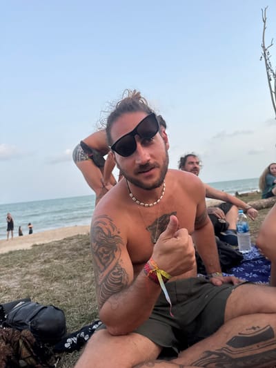 The scene depicts a casual gathering on a beach in Ban Pang Pring, Thailand. In the foreground, a shirtless male subject, appearing to be of adult age, is seated on the ground, looking directly at the camera. He has dark hair styled in a bun, a beard, and wears dark sunglasses. A white shell necklace is around his neck. His right arm is bent, and his hand displays a thumbs-up gesture. Multiple colorful wristbands are visible on his right wrist. Tattoos are present on his left upper arm (a skeletal or flowing figure design), chest, and right thigh (a boat on water). He is wearing dark green shorts. A black bag rests on the ground to his left.
In the midground, to the left of the main subject, another individual is seen bending over, facing away from the camera, wearing dark shorts with a light pattern and a visible leg tattoo. To the right of the main subject, another male is seated, looking towards the right, with a beard. A clear plastic water bottle is on the ground near him, alongside a blue patterned fabric. A partial view of a third seated individual is visible further right.
In the background, the ocean shoreline is visible with gentle waves. Several indistinct figures are present on the sandy beach near the water's edge. The sky is light blue with minimal cloud cover. A thin, bare tree branch or post with small lights or decorations is visible on the far right. The ground transitions from sand near the water to a sparse grassy area where the individuals are seated.