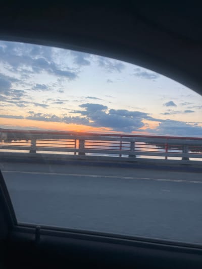 This image captures a view from a vehicle, likely a train, looking out through a window. The dominant scene is a sunset over a body of water, possibly a river, framed by a bridge. The sky is filled with clouds, tinged with the warm hues of orange and pink from the setting sun, transitioning to lighter blues and grays higher up. The bridge structure, with its red railings and vertical supports, is visible on the right side of the frame, casting reflections onto the water below. The water appears calm, mirroring the sky and the bridge.
The foreground is partially obscured by the dark interior of the vehicle, with a curved dark edge suggesting the window frame or a section of the vehicle's interior. On the right side of the image, beyond the bridge, a dark road or pathway is visible, with faint lines suggesting lanes. The overall mood evoked is serene and contemplative, typical of a sunset view during travel. The time of day is clearly sunset, indicated by the colors in the sky. While no people are visible, the perspective suggests they are passengers within the vehicle. There is no visible text in the image.