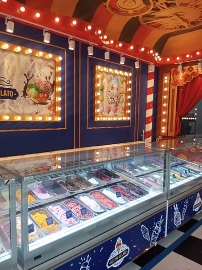 The image depicts the interior of a gelato establishment located in Tân Kỳ Commune, Vietnam, characterized by a prominent circus or carnival theme. In the foreground, two interconnected, glass-enclosed display cases are filled with multiple rectangular stainless steel pans containing various colors of gelato, indicating diverse flavors. Small white labels are visible on some gelato pans. The lower section of the display cases features a blue panel adorned with white line-art illustrations of ice cream cones and the branding "GUSTAV GELATO Italian Gelato." The walls are painted dark blue with gold framing elements. The left wall features a large, rectangular poster depicting gelato scoops in a waffle bowl, bordered by numerous spherical light bulbs. The text "GELATO" is partially visible on this poster. To its right, a smaller, similarly lit poster displays a grid of distinct gelato flavors with the text "Flavors Gelato." The ceiling exhibits a red and dark-toned striped pattern with a central circular ornate detail. A red track lighting system with multiple white spotlights is mounted along the upper wall line, supplemented by additional exposed bulbs. On the far right, a vertical red and white striped column is visible next to red draped curtains, continuing the thematic design. A black and white checkered floor pattern is partially visible in the lower right corner. No individuals are depicted in the image.