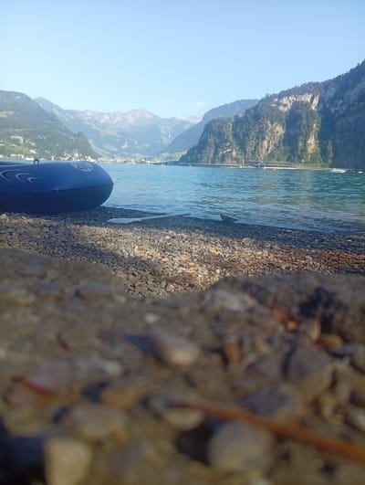 An inflatable blue boat rests on a rocky shore next to a paddle. The boat is partially submerged in a body of water, likely a lake, with mountains in the background. The sky is clear and blue. The scene appears to be in Hergiswil (NW), Switzerland, given the characteristic mountainous landscape of the region.