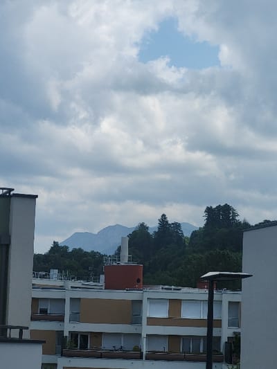 The image shows a view from a residential building in Amphion-les-Bains, France. The sky is overcast with white clouds. In the distance, a mountain range is visible. There are several trees in the foreground, and a streetlight is visible in the lower right corner of the image. The buildings are composed of concrete and brick. They have balconies and windows. There is a bicycle on one of the balconies. The building appears to be constructed in a modern style.