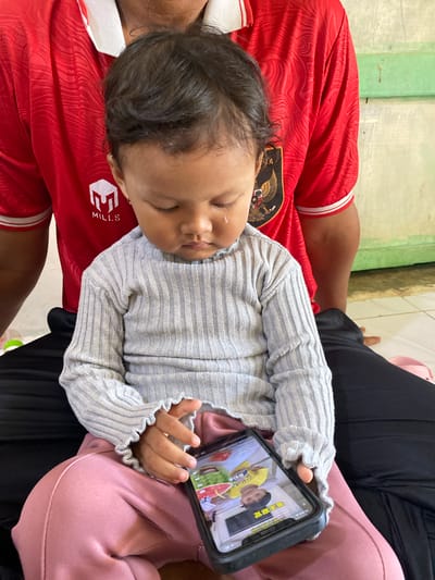 A young child sits on a person's lap, facing downwards and looking at a smartphone. The child wears a grey, ribbed long-sleeved shirt with ruffled cuffs and pink pants. The person they are sitting on wears a red t-shirt with white trim and a logo on the left chest, partially obscured by the child. The smartphone screen displays a video with an Asian child wearing a yellow shirt. The background consists of a textured wall and a green stripe. The scene appears to be indoors, possibly in a home in Tuha Lada, Indonesia.