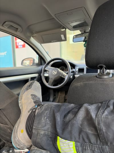 A person wearing grey and black work boots and black work pants with yellow reflective stripes is sitting in the driver's seat of a car. The car's steering wheel has the Opel logo. The rearview mirror and passenger sun visor are visible. The image was likely taken in Ostend, Belgium, as a blue sign with "k wert" is partially visible, which could refer to a location in that city. The pants and shoes appear to be dusty or dirty, suggesting the person has been working.