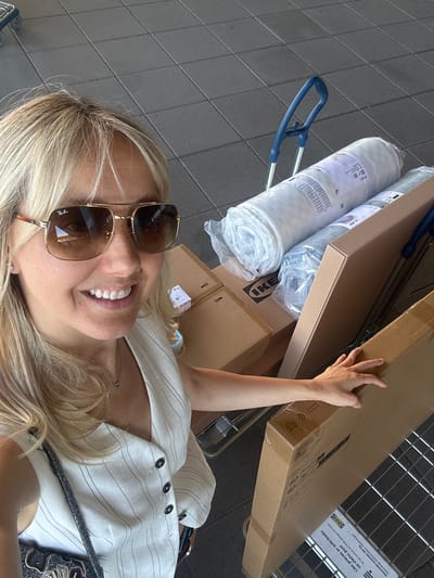 This is a high-angle, selfie-style shot taken outdoors on a tiled surface, likely a sidewalk or parking lot. The central subject is a woman with blonde hair and sunglasses, smiling broadly. She is wearing a white pinstriped top. Behind her, a shopping cart is filled with several large, flat cardboard boxes and two rolled-up items wrapped in white plastic. The IKEA logo is visible on one of the boxes. The hand of the woman is seen reaching towards one of the boxes. The scene suggests a shopping trip, possibly from IKEA, given the prominent branding. The lighting indicates it's daytime. The text visible on the boxes includes "IKEA" and instructions in Latvian, mentioning "Liec preces ar svītrkodu uz vienu pusi" which translates to "Place items with barcode on one side," and "Maksimālā noslodze: 200 kg" meaning "Maximum load: 200 kg." The presence of Latvian text strongly suggests the location is in Latvia, with Rīga being a likely urban setting for such a purchase.