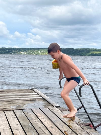 A young boy wearing dark blue swim trunks and yellow arm floaties is stepping off a wooden dock into a body of water, holding onto a metal ladder. The boy is facing to the right of the frame, looking down. The dock is made of weathered wooden planks. The water is dark and appears rippled. In the background, a tree-lined shore is visible with a few buildings scattered along it. The sky is overcast with clouds. This scene is located on the Sylva River in the city of Sylva, Russia.