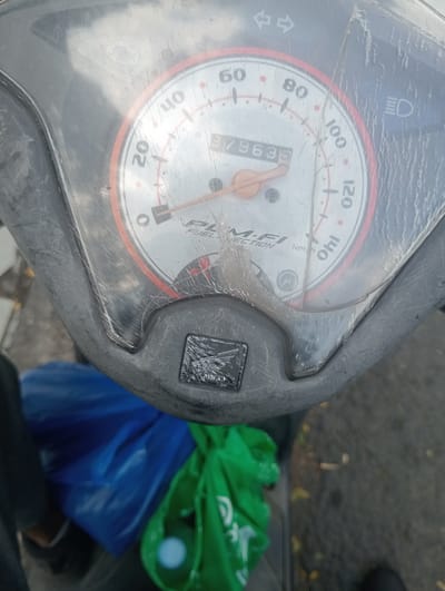 The image shows a close-up of a motorcycle's instrument panel. The speedometer reads 79635, and the panel includes indicators for fuel and headlights. The instrument panel is covered by a scratched plastic shield. Below the instrument panel, a Honda logo is visible. In the background, out of focus, are two plastic bags, one blue and one green. The ground beneath appears to be asphalt. The context of Surabaya, East Java, is not directly evident from the image itself but is provided as additional information.