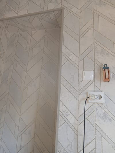The image shows a wall with geometric patterned wallpaper in what appears to be an interior space. There are two electrical outlets and one light switch visible on the wall. A black cable is plugged into the lower electrical outlet. The wallpaper has a light color with a pattern of intersecting lines and textured sections that resemble marble. The overall scene is indoors, and no specific outdoor elements are visible. The location is identified as Kushkayragach, Uzbekistan.