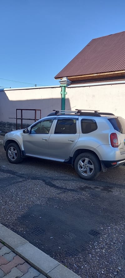 A silver Dacia Duster is parked on a gravel surface next to a building with a brown metal roof. The car is viewed from the side, facing left. A green pole with a decorative white top stands near the car. The sky is clear and blue. The ground is a mix of gravel and some dark patches, possibly asphalt or oil stains. A paved sidewalk is visible in the foreground. The scene appears to be outdoors in a residential or industrial area.