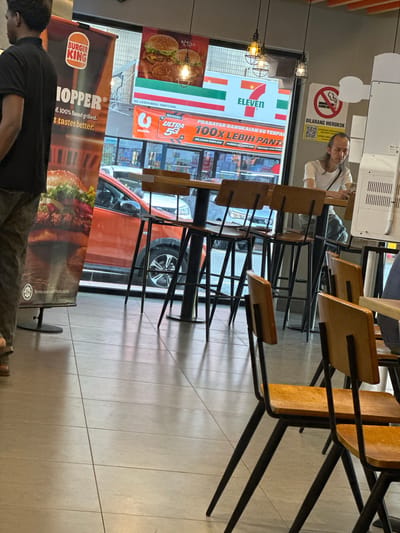 The image displays the interior of a Burger King restaurant in Subang Jaya City Council, Malaysia, with a large glass window offering a view of a commercial street.
In the left foreground, a person is partially visible from the back, wearing a dark short-sleeved top and light-colored trousers. In the mid-ground right, a man with long dark hair, dressed in a white short-sleeved t-shirt and light-colored trousers, is seated at a high-top table, focused on a smartphone in his hands. A dark strap crosses his chest.
The restaurant interior features light-toned square floor tiles with dark grout lines. High-top tables with dark metal bases and wooden tops are arranged with matching high-back bar stools, featuring dark metal frames and light brown wooden seats and backrests. Three industrial-style pendant lights with wire cages and exposed bulbs hang from the ceiling. A tall, white vertical structure with ventilation grilles is present on the far right. A white rectangular sign on the right wall prominently displays "DILARANG MEROKOK" (No Smoking) with a crossed-out cigarette symbol and two QR codes.
On the left, a large Burger King banner is visible, partially obscured, displaying "BURGER KING" and text including "HOPPER," "100% flamed-grilled," and "tastes better." A "Halal" logo and "Terms and conditions apply" are located at the bottom of this banner.
Through the window, an orange SUV is parked, partially obscuring the street view, with its front passenger side wheel and fender clearly visible. Across the street, a prominent "7 ELEVEN" sign, characterized by its red, green, and white stripes, is visible. Below the "7 ELEVEN" sign, a large advertisement banner for "U mobile" (partially visible logo) promotes "ULTRA 5G" and "100X LEBIH PANTAS" (100x Faster). Above the "7 ELEVEN" sign, a promotional poster features burgers and fried chicken, with "NO. LESEN MBSJ: 186871738B" printed on a grey strip.