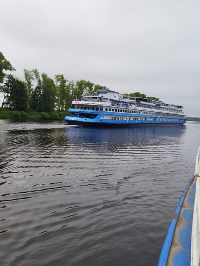 A long, blue and white cruise ship travels on a wide, dark river under an overcast sky. The ship is positioned in the middle of the frame, angled away from the viewer and towards the right. It has multiple decks with numerous windows. Green trees and foliage line the riverbank on the left side of the image. In the foreground, the edge of another boat, painted blue and white, is visible on the right. The water is rippled, creating a textured surface. The scene appears to be in Faliukovo, Russia, likely on a river cruise route.