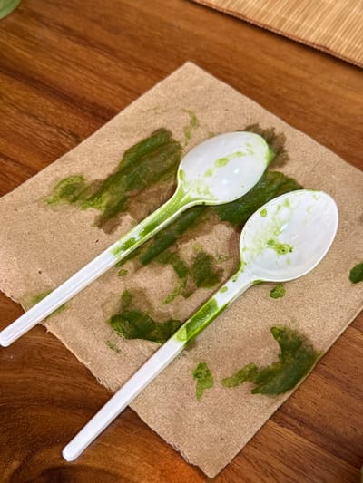 Two white plastic spoons, heavily stained with a vibrant green, somewhat pasty substance, are depicted resting diagonally on a light brown paper napkin. The napkin is also marked with numerous splashes and smears of the same green material, indicating recent use during a meal. The spoons are positioned with their bowls facing upwards, showing remnants of the green food item both inside the bowls and along their handles.

The setting appears to be a casual dining environment. The napkin and spoons are placed on a warm-toned wooden table, which features a visible grain, covering much of the background. A textured mat, possibly made of woven bamboo or wood strips, is partially visible in the upper left corner of the frame. The image is captured from a high-angle perspective, looking down onto the table.

The scene suggests the conclusion of a meal where a green-colored dish, possibly a sauce, soup, or dessert, was consumed. There is no discernible activity taking place, only the aftermath of dining. No visible text is present in the image. The photo depicts an everyday moment from what could be a restaurant or cafe in Guadalajara, Mexico.