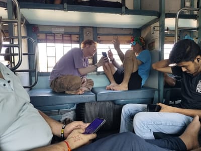 The image shows the interior of a passenger train compartment in Kelva, India. Four passengers are visible. Two Caucasian men, one wearing a purple shirt and khaki pants and the other in a blue shirt, black shorts, and a colorful headband, are sitting opposite each other on a blue bench, both looking at their cell phones. The first man is fair-skinned, with red hair. The second man is wearing sunglasses. Two Indian men are also present. One sits to the side, listening to music and using his phone; the other is sitting in the foreground, also looking at his phone. The first man is wearing a black T-shirt, and the second man is wearing a white shirt. In the foreground, the man's arm has a tattoo. Bags are stored under the bench. The upper berth of the compartment is visible, with bedding stored on it. The windows are letting in bright sunlight.