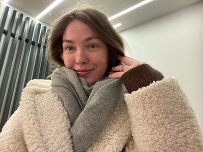 The image displays a close-up indoor portrait of a young adult female located in Salar'yevo, Russia. She has light skin, blue eyes, and light brown, wavy hair. Her expression is a faint smile, with slightly parted, glossy lips. She is dressed in a cream-colored, textured, fleece-like jacket and a light grey scarf wrapped around her neck. Her right hand is raised, with fingers touching her hair near her right ear; a thin, gold-toned ring is visible on her right ring finger. The cuff of a brown, ribbed, possibly sparkly garment extends from beneath the jacket sleeve on her right wrist. Her left arm supports her head, resting against her cheek. The background consists of a section of a white wall to the right and a decorative panel to the left, featuring vertical, dark gray rectangular slats. An overhead linear light fixture is visible along the white ceiling.