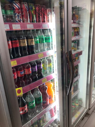 This is a medium shot, eye-level, indoor photograph of a brightly lit beverage refrigerator display. The refrigerator, located in Putat, Indonesia, is filled with various bottled and canned drinks. The left side of the refrigerator is filled with soda bottles, primarily Coca-Cola and Fanta, in red, orange, and clear plastic bottles. There are also several bottles of Sprite, a clear carbonated beverage, with green labeling. Above the soda bottles, on a top shelf, are several cans of Coca-Cola, with red and white designs, and other soda cans with various colorful designs. The right side of the refrigerator contains more canned drinks, including what appears to be Fanta and other brands with labels in Indonesian. The refrigerator doors are glass, allowing a clear view of the products inside. The overall environment appears to be a small convenience store or a similar retail establishment.