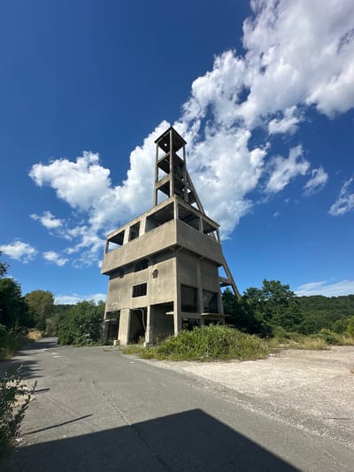 An abandoned concrete structure, likely a mine shaft headframe, stands under a bright blue sky with scattered white clouds. The structure is multi-tiered and appears to be in a state of disrepair, with open doorways and missing sections. Vegetation, including trees and bushes, surrounds the base of the structure and encroaches on a cracked asphalt road leading away from it. The scene is situated in a rural or semi-rural setting, with a wooded hillside visible in the background. The lighting suggests it is daytime. There are no people or animals present in the image. The location is identified as Rosen, Bulgaria.