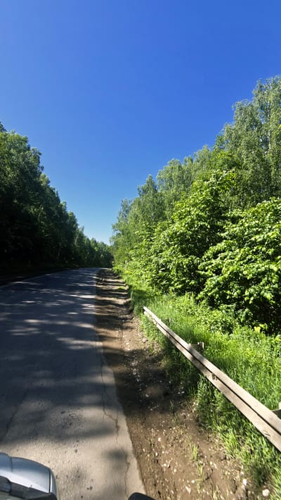 The image shows a rural road lined with lush green trees on both sides. The road appears to be paved with asphalt and has some cracks and shadows from the overhead foliage. The sky is a clear, bright blue. To the right of the road, there is a wooden guardrail running along the edge, with tall green grass and some dirt. The scene suggests a sunny day in a natural setting. The location is identified as Актюбинский, Russia. The image appears to be taken from a moving vehicle, as indicated by the slight blur and the perspective of the road. There are no people or animals visible in the image.