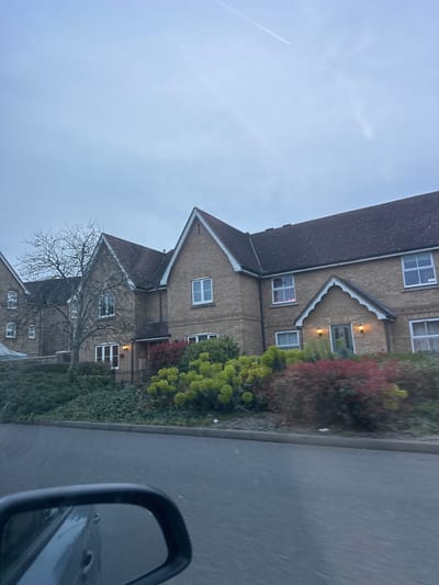 The image depicts a multi-story residential or institutional building located in the city of Gillingham, United Kingdom, viewed from a vehicle on an adjacent road under an overcast sky. The sky is a pale grey, with a faint contrail visible in the upper right quadrant.
The building is constructed primarily of light brown or buff brickwork, featuring multiple gabled sections with dark grey tiled roofs. White-framed windows are visible across all visible floors, including some within the gabled dormers. Architectural details include stepped brickwork below several windows and along the edges of the gables. One upper-story window on the right side of the main gabled section emits a distinct pink light from within.
Two illuminated entrances are visible. The most prominent entrance, positioned centrally, features a decorative gabled porch with light green double doors and two active wall-mounted sconces casting a warm light. Another entrance is visible further to the left, also with an active outdoor light fixture. A bare deciduous tree stands in front of the left-most section of the building, suggesting a cold season.
The foreground consists of a dark grey asphalt road, separated from the building by a grey curb and a landscaped area. The landscaping features a dense array of shrubs, including dark green ground cover, brighter lime-green bushes, and reddish-brown foliage, indicating deliberate planting and maintenance. Small white fragments, possibly litter, are visible on the curb and the edge of the landscaped area. The lower-left corner of the image includes the black casing and reflective surface of a vehicle's side mirror, indicating the photograph was taken from inside a moving or stationary vehicle. No people or active interactions are visible within the scene.