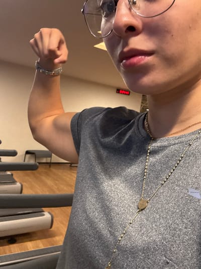A person in a gray shirt flexes their arm. The individual is wearing glasses and necklaces. Their hand is clenched into a fist. Part of their face is visible, including their nose and lips. The background features a wall, a digital display showing "DOWN," and exercise equipment, possibly treadmills, with gray supports and white consoles. The floor appears to be wooden.