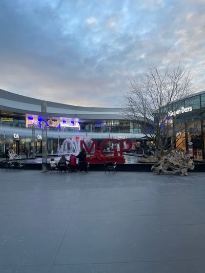 The scene depicts an outdoor plaza within a modern shopping center, specifically identified as being located in Plaisir, France. The foreground is a wide, flat grey paved surface. In the mid-ground, a rectangular water feature contains the prominent red letters "MGP," which are internally illuminated and adorned with red floral or decorative elements atop the 'M' and 'G'. Water jets are active within the pool surrounding these letters. To the right of the water feature, a large, multi-branched leafless tree stands, with a gnarled wooden sculpture resting on the pavement near its base.

Four individuals are seated at a small black table with black chairs to the immediate left of the "MGP" sign; a red stroller or pram is positioned next to the leftmost person. Numerous other unoccupied black chairs are arranged in rows across the paved area. The background features a multi-level, curved architectural structure with a grey and white banded facade, housing various storefronts. Visible store signage includes "OLD WILD WEST," "C&A," "CASHVILLE," and "Häagen-Dazs." Blue decorative lighting is integrated into the building's exterior and display windows. Multiple indistinct human figures are visible through the glass facades of the stores. The sky overhead is overcast with light blue-grey cloud cover.