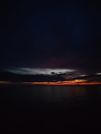 The image depicts a seascape at twilight with a dramatic sunset. The sky is dominated by dark, heavy clouds with streaks of deep purple and hints of lighter gray. Near the horizon, a band of intense orange and red light glows from the setting sun, illuminating the undersides of the clouds. Below the sky, a vast expanse of dark, calm water stretches to the horizon, reflecting the muted colors of the sky. The overall scene is somber and atmospheric, with the darkness of the night gradually consuming the last light of day. The location is specified as Kvartsitnyi, Russia, suggesting this is a view of the sea from that coastal area. There are no people, specific objects, or actions visible in the frame, focusing solely on the natural landscape.