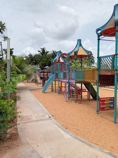 The image shows a colorful playground structure on a sand surface, likely a playground in Chopdem, India. The structure is made of metal and plastic, with slides, climbing features, and interactive elements. Palm trees and other vegetation are visible in the background, suggesting a tropical climate. A stone walkway borders the playground. The sky is overcast. No people are visible.