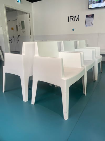 This is an interior shot of a modern, empty waiting room in a medical facility, likely in Clermont-Ferrand, France, based on the context. The photograph captures a long row of empty, white, angular chairs with a contemporary, sculptural design, arranged along a wall. The chairs are made of a molded material and show some minor scuff marks.
The room features a clean, two-tone color scheme. The floor and the wall directly behind the chairs are a distinct blue-green. The opposite wall, to the left of the frame, is white, as is the ceiling.
On the white wall, large black letters spell "IRM" (Imagerie par Résonance Magnétique / MRI). Above this, a small blue circular dot is visible. Further up, a white poster provides information, with text partially visible, including "RADIOLOGIE POUR TOUS ET PAR..." (Radiology for all and by...), mentioning "rendez-vous" (appointment), "clichés (radiographie)" (X-rays), "Scanner," and "CD avec images." A QR code is also present on the poster. To the left of the poster, part of a screen with "S3" visible is mounted on the wall. A green emergency exit sign is also seen on the far left.
An opening in the white wall reveals an adjacent area with white doors. One door is clearly marked "SALLE D'ATTENTE" (Waiting Room) and "2," accompanied by universal warning symbols (e.g., no food/drink, no mobile phones). The overall impression is one of a bright, well-lit, and professionally managed medical environment designed for patient comfort.