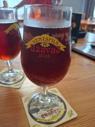 A medium shot captures two glasses of dark beer on a wooden table. The glass in the foreground, which is larger and in focus, displays a yellow ribbon-like label with "VENTSPILS" written in black at the top, followed by "ANNO 1994" and "UŽAVAS" in red script. Below this, "alus" is written in smaller red script, and then "Ģimenes Alus Darītava" in grey. The beer has a frothy head and condensation on the glass. The second glass, out of focus to the left, shows a similar label partially obscured. Coasters with similar branding are beneath each glass. The background is blurred, suggesting an indoor setting with people and furniture, possibly a pub or restaurant in Lēdmane, Latvia, given the branding on the glasses.