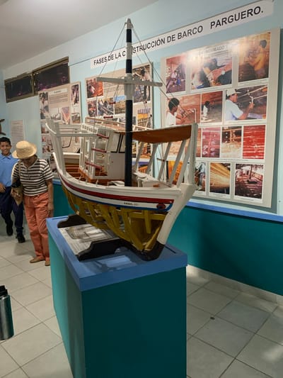 The image depicts an indoor exhibit featuring a model of a boat. The model is placed on a blue pedestal in the foreground. Behind the model, a large poster displays photographs illustrating the "Phases of Boat Construction" with text in Spanish. The walls are painted in a light blue and teal color. Two individuals are visible in the background: a young male wearing a blue shirt and a woman in a straw hat, striped shirt, and orange pants. They appear to be observing the exhibit. The floor is tiled with light-colored squares. The overall scene suggests a museum or exhibition space. The mention of Boca del Río, Venezuela, as the location is based on external knowledge and is not directly inferable from the visual elements alone.