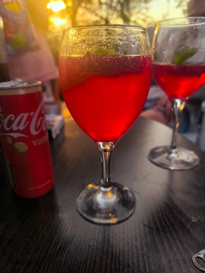 The image shows a close-up of two glasses of red beverages and a can of Coca-Cola on a dark wooden table. The beverage in the foreground is in a wine glass and has a garnish of mint leaves. Bubbles are visible within the liquid, indicating it is carbonated. The glass has condensation on its exterior. A second, similar glass of red beverage is visible in the background to the right, also containing mint leaves. To the left of the foreground glass is a red Coca-Cola can, with visible branding. The background is blurred and shows warm, golden light, possibly from a sunset, and indistinct figures of people. The setting appears to be an outdoor area, potentially a restaurant or bar. There is no specific visual information to identify the location as Almere, Netherlands.