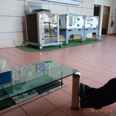 The image shows a room with tiled floors. There is a glass-top coffee table with silver legs in the foreground, and a pair of feet, wearing dark shoes, is visible in the right corner. The room contains three pieces of industrial equipment, each mounted on a metal platform with wheels. Two are rectangular with metal grilles and are labeled “GARUDA”. One has a large glass door and is labeled “GARUDA”. The equipment is in the center of the room and is surrounded by green turf. The room appears to be part of an office or industrial building. The location, based on coordinates -6.338, 107.139, is in the city of Sukaresmi, Indonesia.